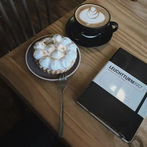 a cup of latte art coffee, lemon tart, and a black leuchtturm1917 journal on a wooden table