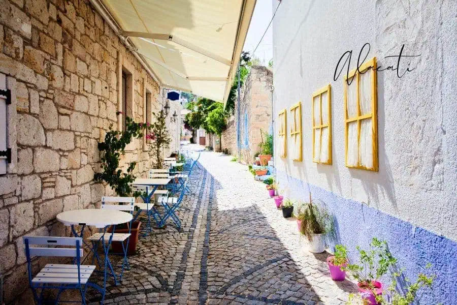 picturesque narrow alley in alaçatı with rustic stone buildings, blue and white chairs, and flower pots decorating the walls