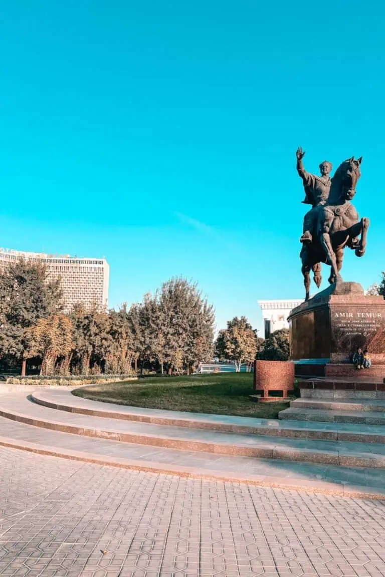 amir temur statue in tashkent, uzbekistan, showing the iconic equestrian monument with hotel uzbekistan in the background, surrounded by trees under a bright blue sky