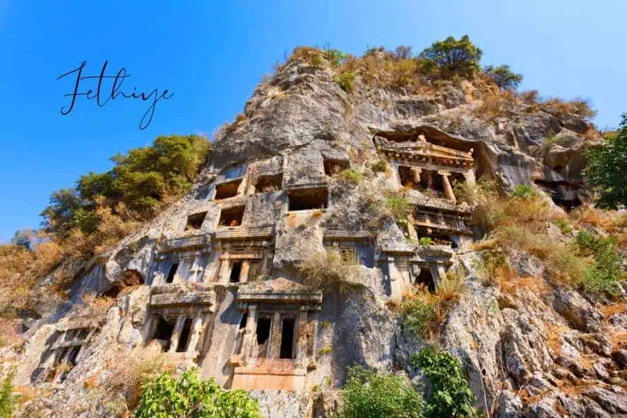 ancient lycian amyntas rock tombs carved into a rugged cliffside in fethiye, surrounded by dry vegetation under a clear blue sky