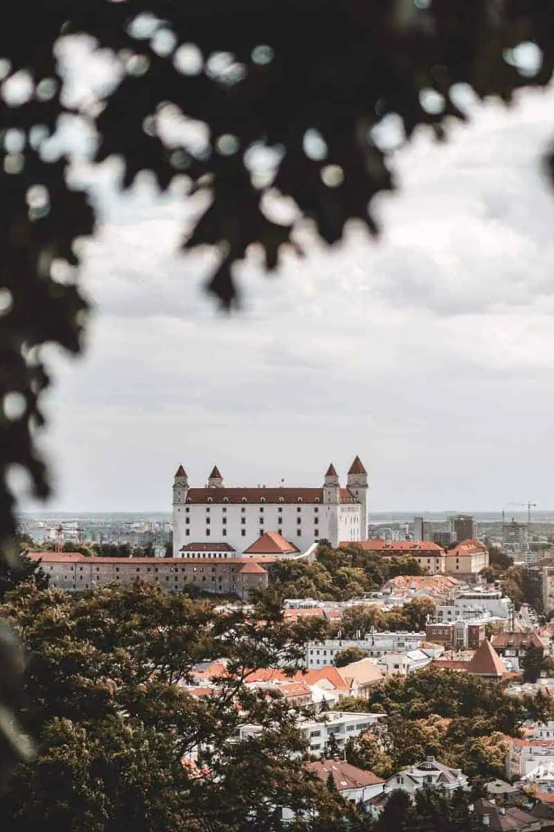 distant view of the white Bratislava Castle with its red rooftops and four corner towers, perched on a hill overlooking the city of Bratislava, Slovakia, framed by leafy branches in the foreground