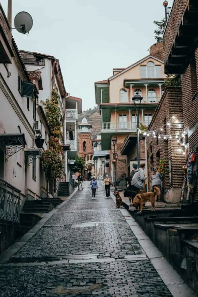 A cobbled street in Tbilisi’s Old Town with colorful balconies, vintage-style lanterns, and a few locals walking past sleepy street dogs