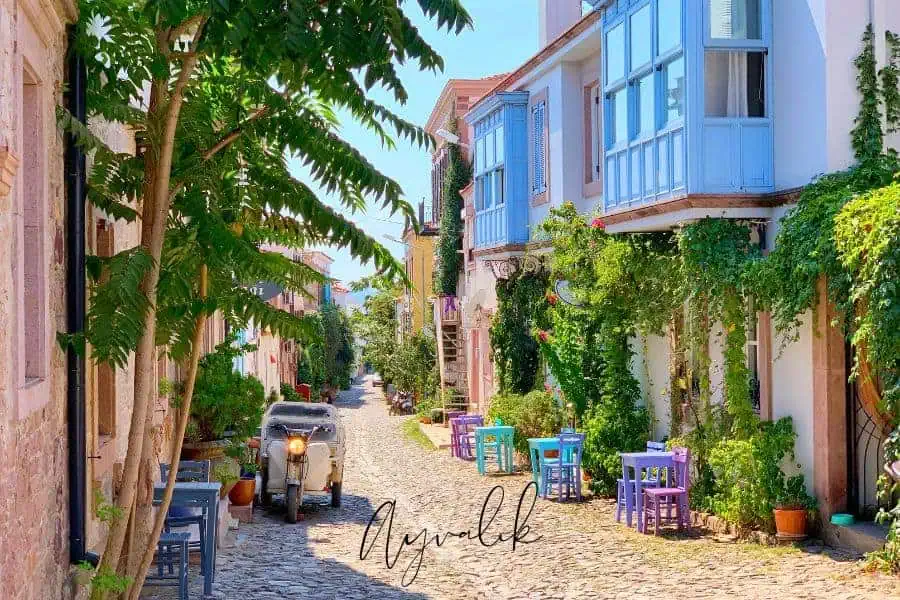 charming cobblestone street in cunda island, ayvalık, one of the most beautiful places in turkey, lined with colorful chairs, lush greenery, and pastel-colored historic houses with blue shutters