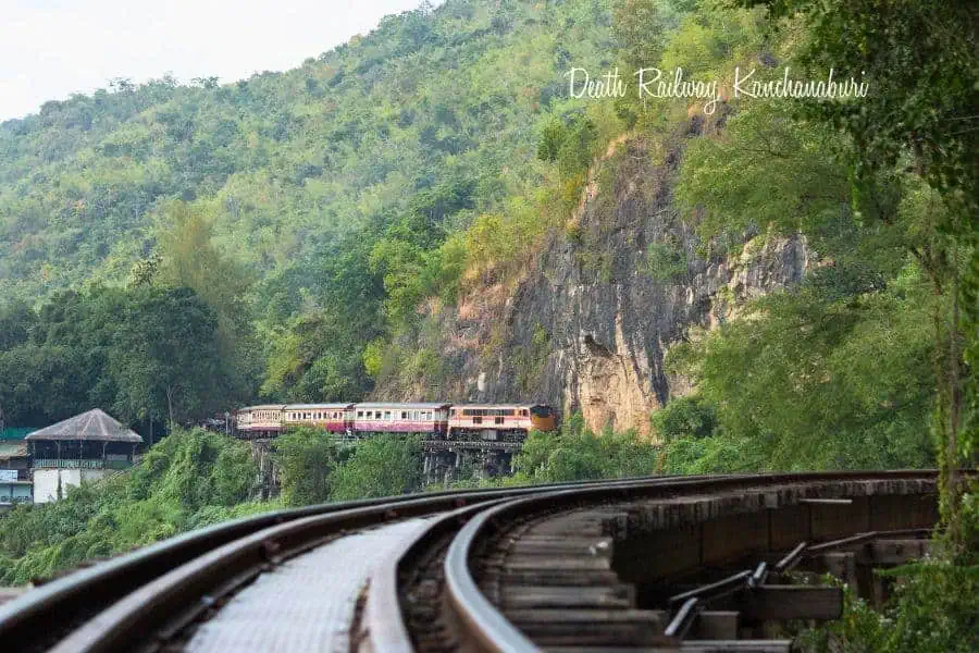 train traveling along the historic death railway in kanchanaburi, thailand, passing through lush green hills and a steep rocky cliffside