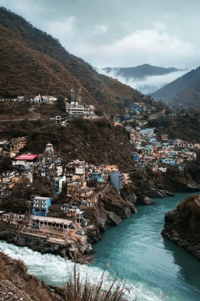  devprayag, the sacred confluence where the rivers alaknanda and bhagirathi meet to form the holy ganga, surrounded by hillside homes and lush mountains under a misty sky