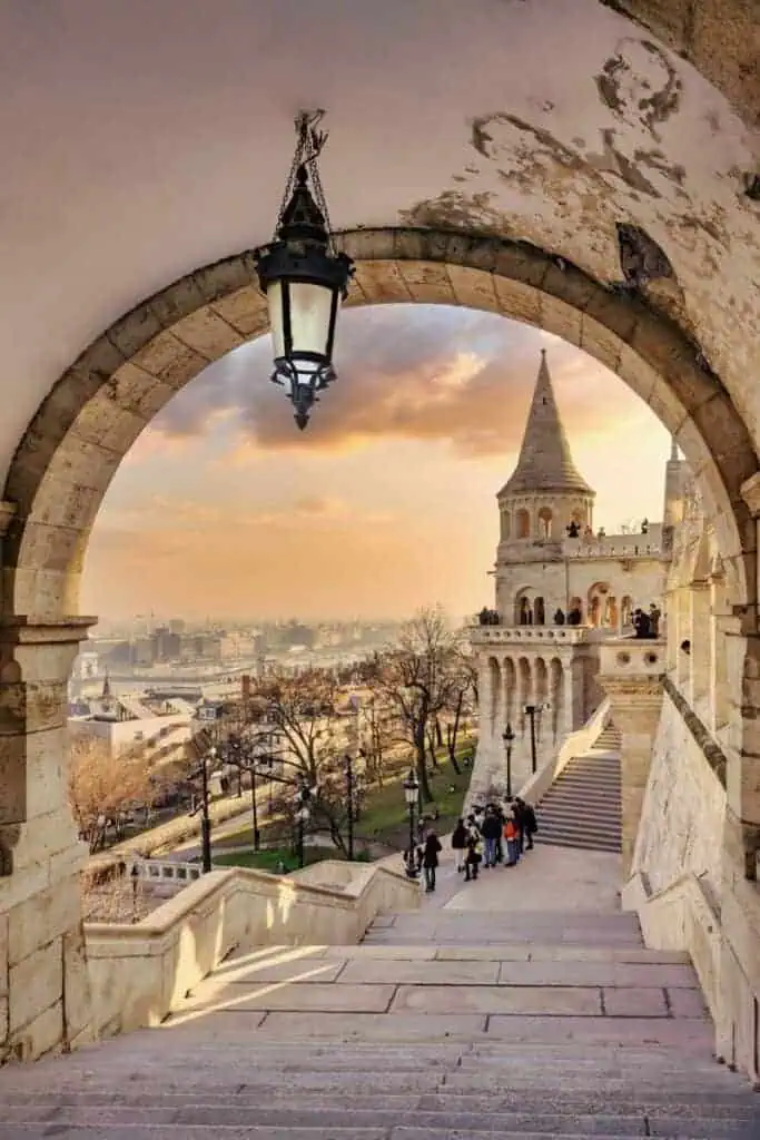 arched viewpoint framing the fisherman’s bastion in budapest, hungary, with people walking along the steps at sunset overlooking the danube river and cityscape