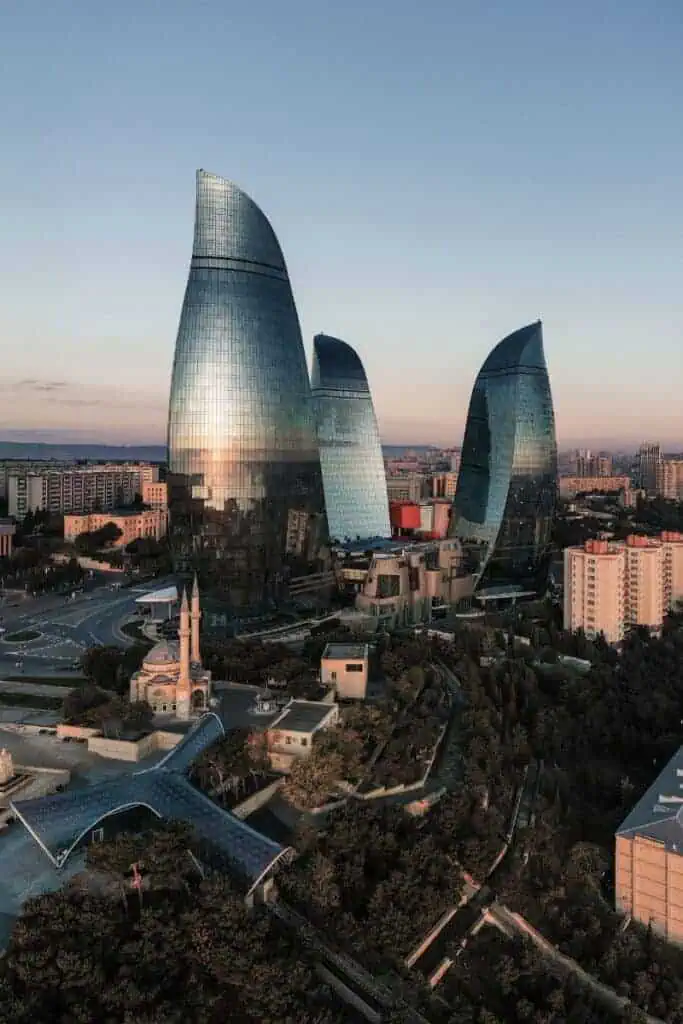 flame towers in baku, azerbaijan, reflecting the soft light of sunset over the cityscape, surrounded by modern and historic buildings with tree-lined streets below