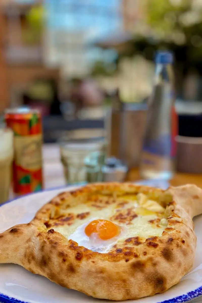 georgian khachapuri filled with melted cheese and topped with a runny egg yolk served on a white plate in an outdoor cafe setting in tbilisi