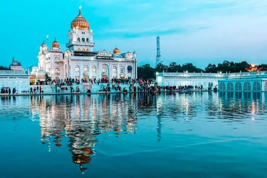 evening view of gurudwara bangla sahib with golden domes glowing and reflecting in the sacred pond, surrounded by devotees and city skyline in the backdrop