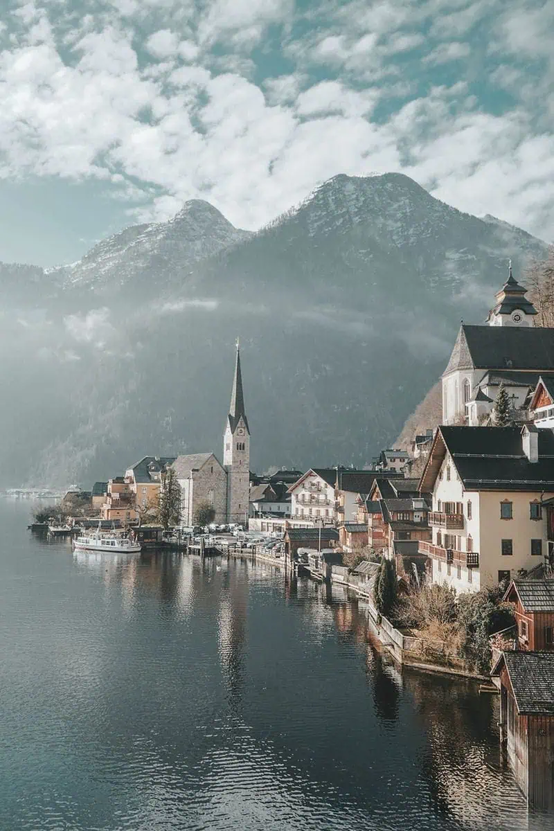 scenic view of Hallstatt village in Austria with lake, church steeple, alpine houses, and misty mountains in the background