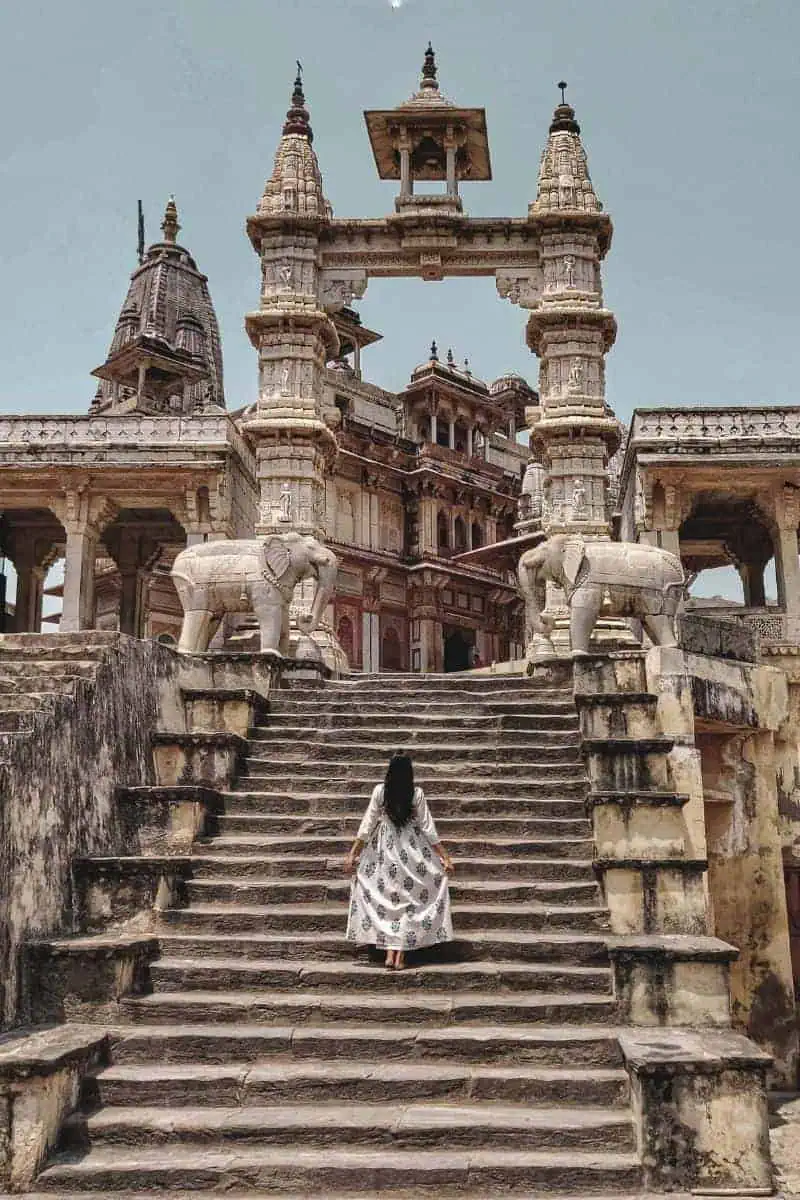 a woman in a white dress walking up the ancient stone steps of a historic temple adorned with intricate carvings and elephant statues, under a clear sky