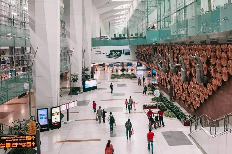 inside view of delhi’s indira gandhi international airport terminal, with passengers walking across the spacious, modern concourse, and the iconic hand mudra wall installation displayed prominently on the right side