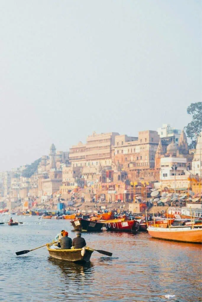 boatmen rowing on the ganges river with the ancient ghats of varanasi in the background, surrounded by colorful boats, historic temples, and buildings under a hazy sky
