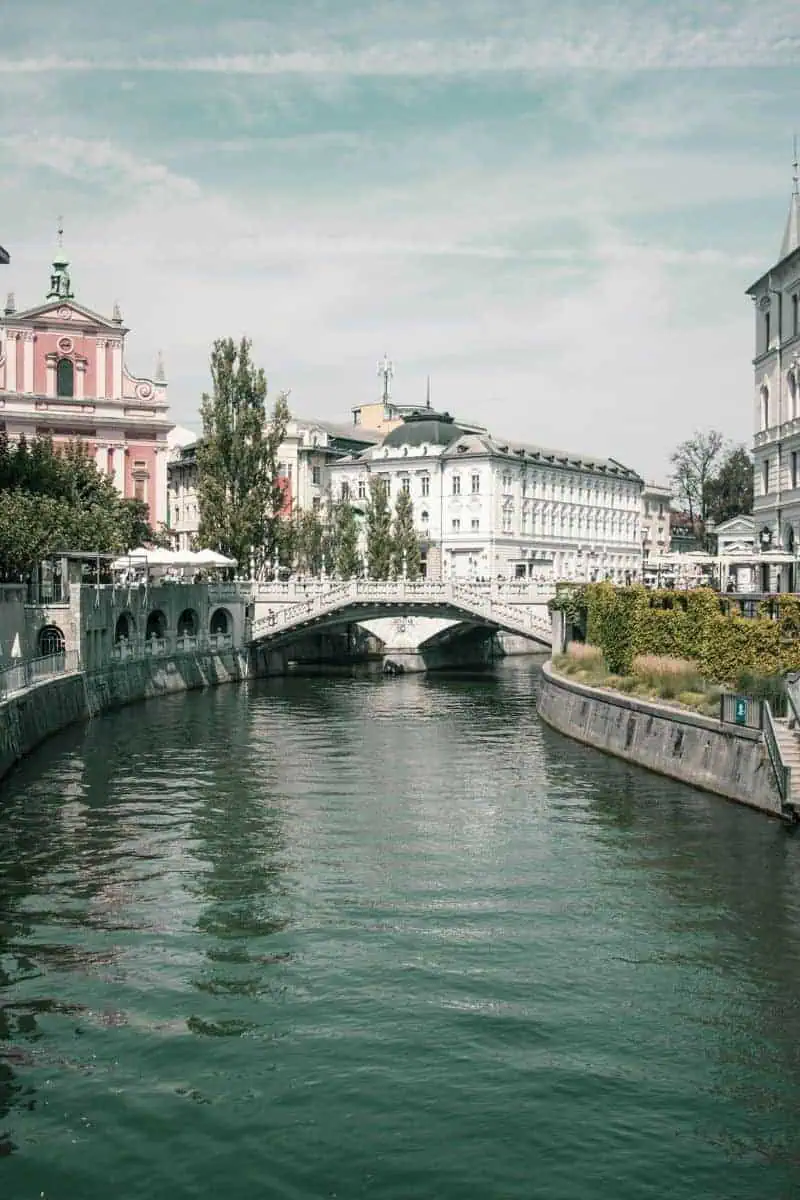view of the Triple Bridge in Ljubljana, Slovenia, spanning the Ljubljanica River with historic white buildings and the pink Franciscan Church of the Annunciation in the background under a partly cloudy sky