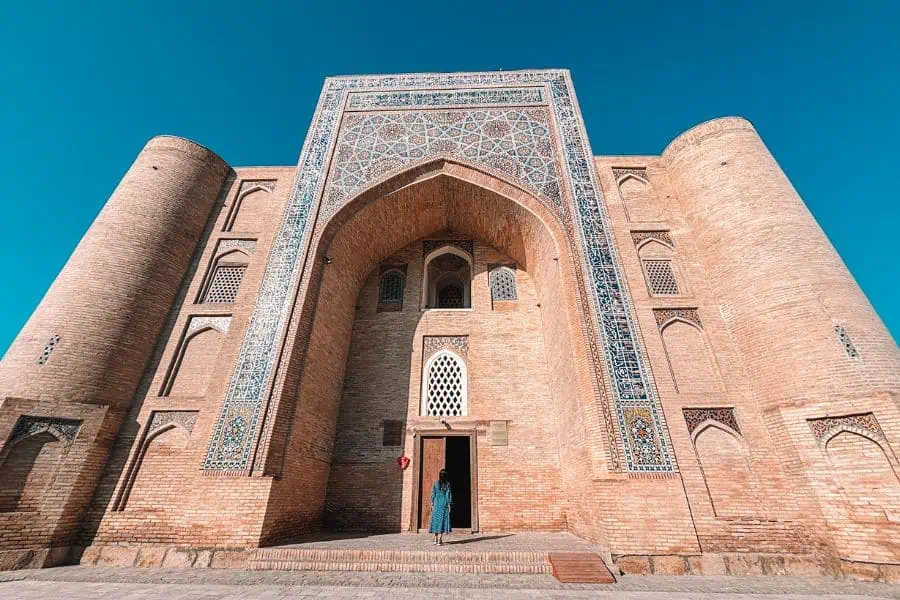 me standing at the entrance of nadir devonbegi madrasa in bukhara, framed by a grand brick iwan with intricate tilework and twin cylindrical towers under a clear blue sky