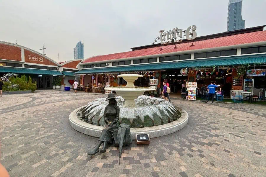 an open-air market scene at asiatique the riverfront in bangkok thailand featuring a central fountain with a bronze statue of a seated figure surrounded by shops with signage and pedestrians strolling by