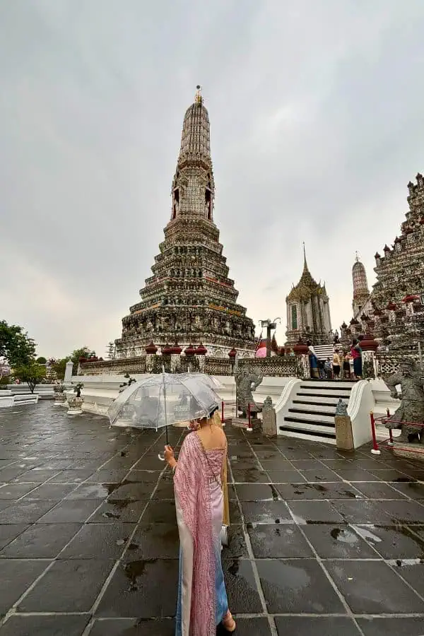 a girl in a traditional thai costume holding a clear umbrella stands facing the ornate spire or prang of wat arun, the temple of dawn on a cloudy day in bangkok, thailand