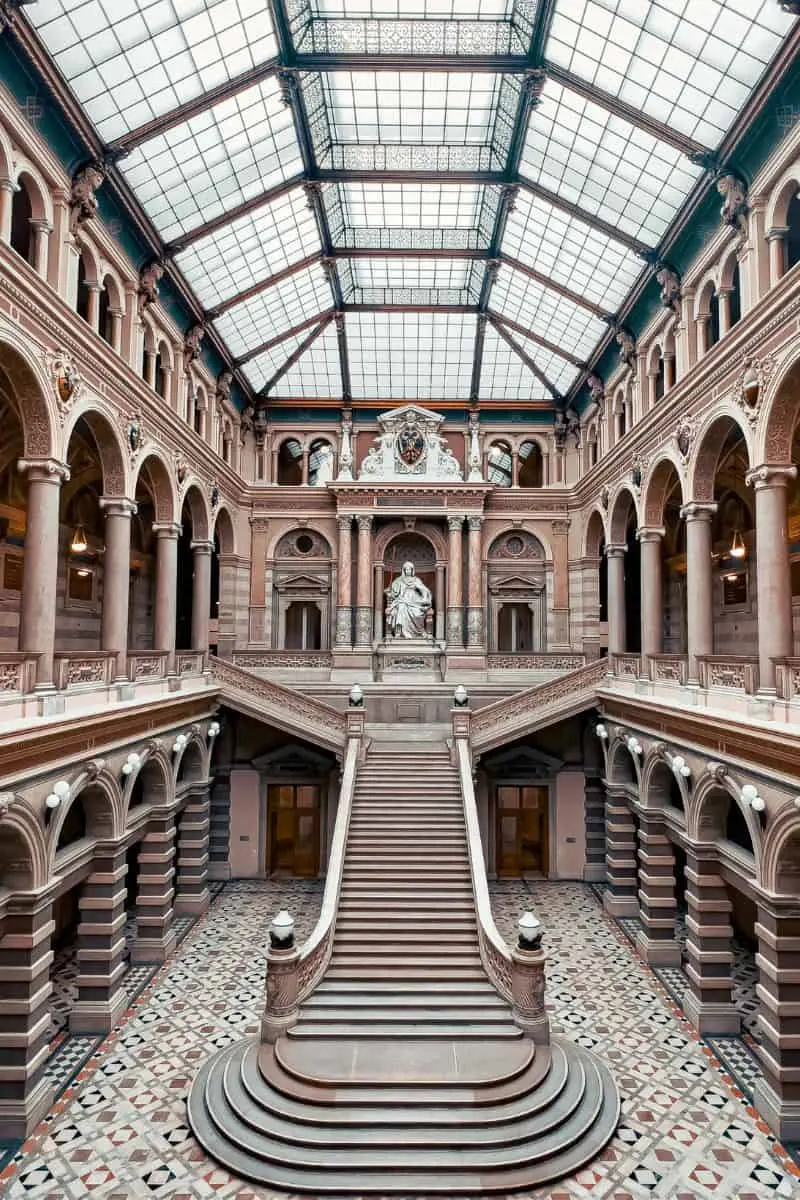 grand staircase inside the palais of justice in vienna, austria, featuring ornate arches, marble columns, a patterned floor, and a central statue beneath a striking glass ceiling