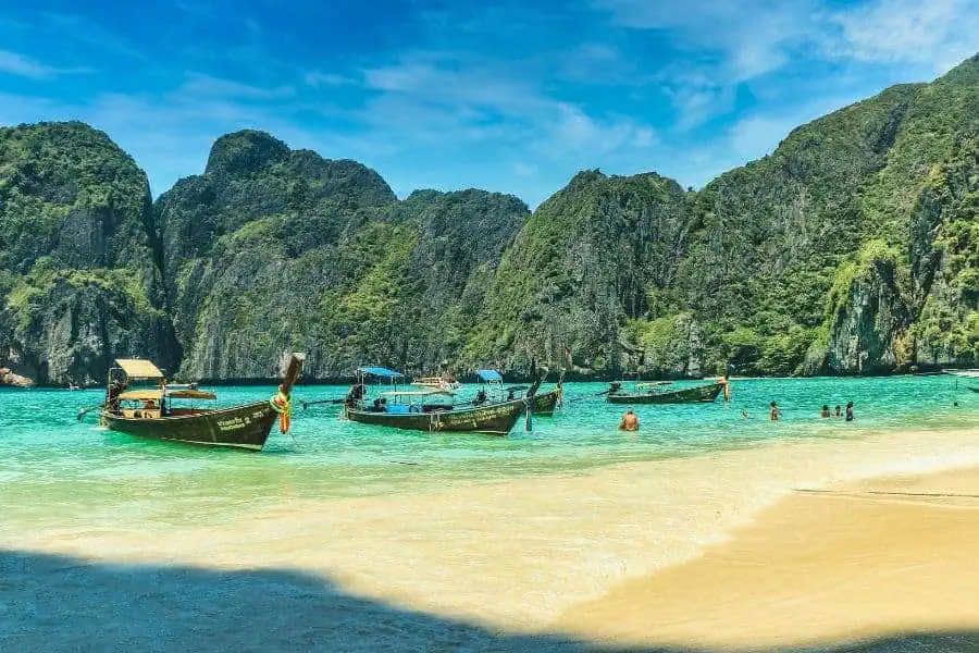 longtail boats floating on turquoise waters at maya bay, phuket, surrounded by towering limestone cliffs with a sandy beach in the foreground