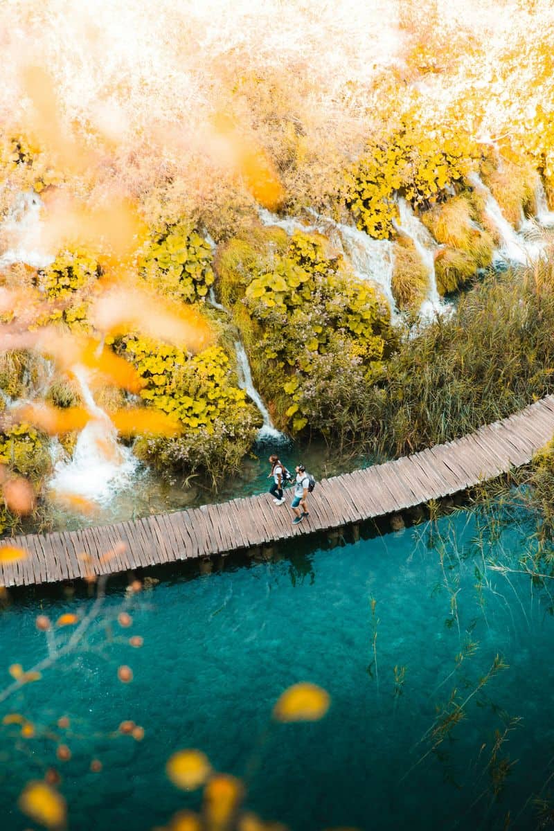 people walking on a wooden boardwalk over turquoise water at plitvice lakes national park, croatia, surrounded by autumn foliage and small cascading waterfalls