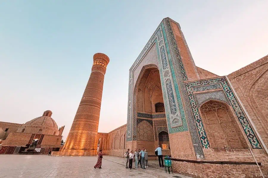 poi kalyan complex in bukhara at sunset with people walking near the illuminated minaret and turquoise-tiled madrasa under a clear sky