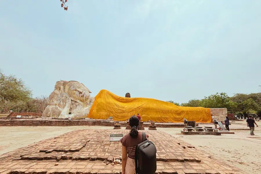 mini-me walking towards the massive reclining buddha at wat lokayasutharam in ayutthaya, thailand, draped in a bright yellow cloth, with ancient brick ruins surrounding the site