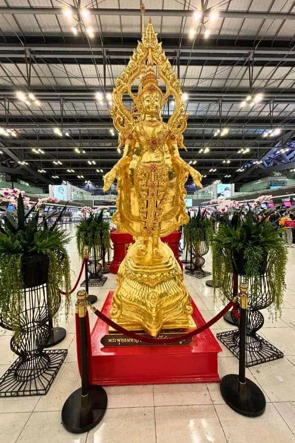 golden statue of a thai deity on a red platform, surrounded by green plants and velvet ropes on suvarnabhumi airport in bangkok thailand