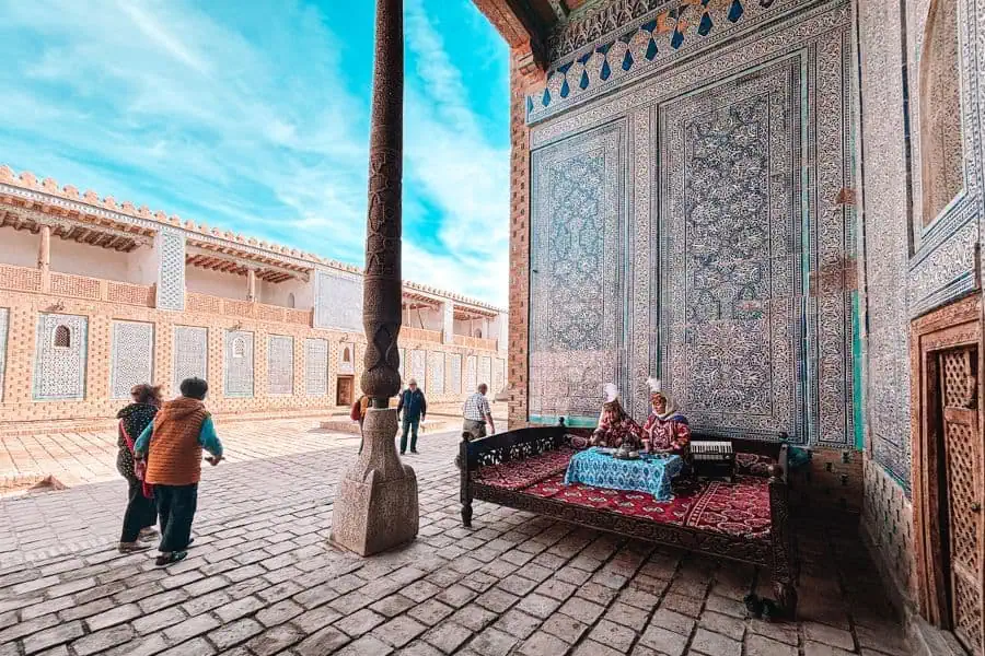 women seated on a traditional wooden daybed inside tosh hovli palace in khiva, surrounded by ornate blue tilework as visitors walk through the palace courtyard