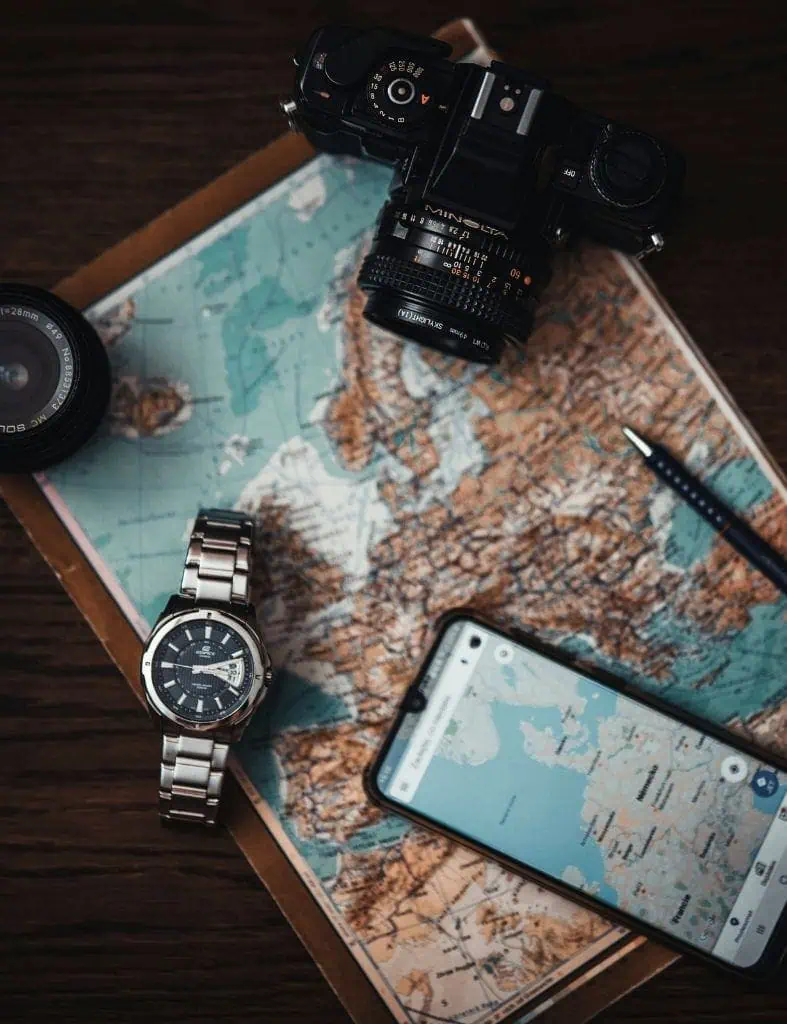 flatlay of a vintage camera, wristwatch, smartphone with maps app open, and a pen on a world map — travel essentials for planning and navigation