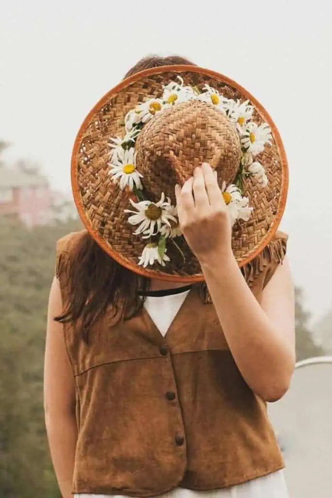 a woman wearing a brown suede vest and holding a straw hat decorated with daisies in front of her face, standing outdoors on a misty day
