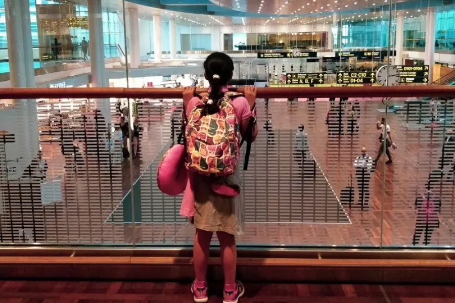 A young girl with a colourful backpack and travel pillow peering over an airport railing, ready for the start of a family Scandinavia trip
