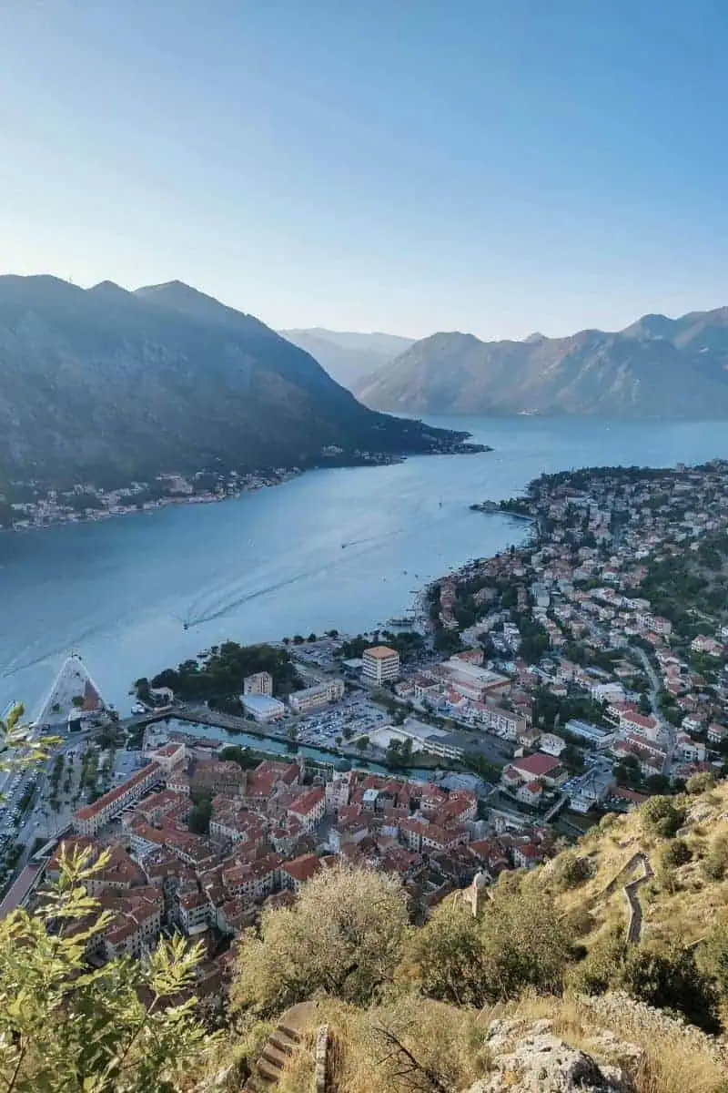  aerial view of the Bay of Kotor in Montenegro, showcasing the red-roofed old town of Kotor, surrounded by dramatic mountains and calm blue waters, with winding roads and boats visible in the bay