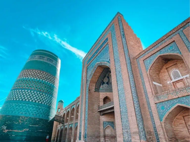 kalta minor minaret in khiva with its short turquoise-tiled cylindrical tower beside the mohammed amin khan madrasa, showing tall brick arches, blue geometric tilework, and a bright clear sky overhead