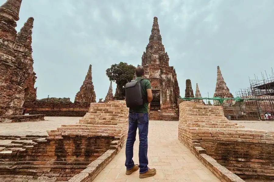 my husband standing with a backpack, admiring the ancient ruins of wat chaiwatthanaram in ayutthaya, thailand, surrounded by towering brick prangs and weathered temple structures