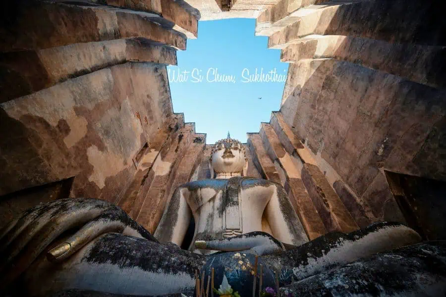 massive seated buddha statue at wat si chum in sukhothai, thailand, framed by towering ancient stone walls with a bright blue sky above