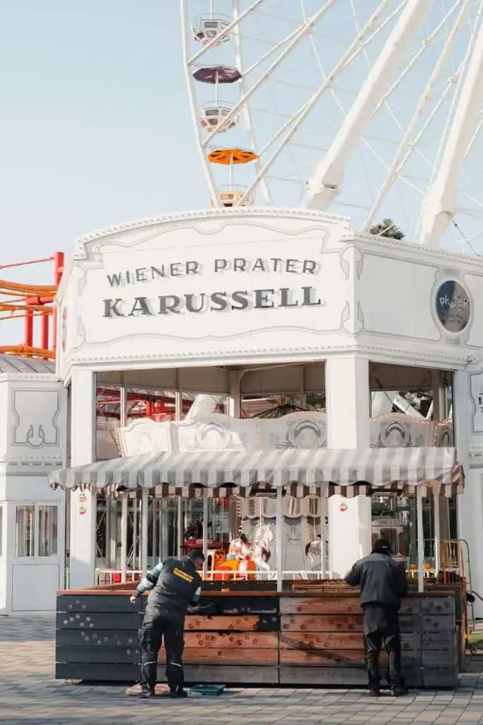 workers painting a wooden counter in front of Wiener Prater Karussell, a carousel at an amusement park with a Ferris wheel in the background