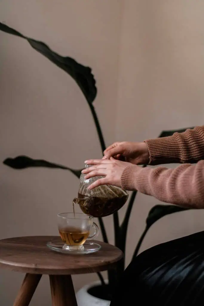 a woman in a brown sweater pouring herbal tea from a glass teapot into a clear cup on a wooden stool, with a leafy plant in the background