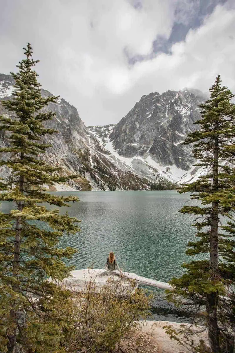 woman sitting on a rock by a turquoise alpine lake surrounded by snow-capped mountains and pine trees under a cloudy sky