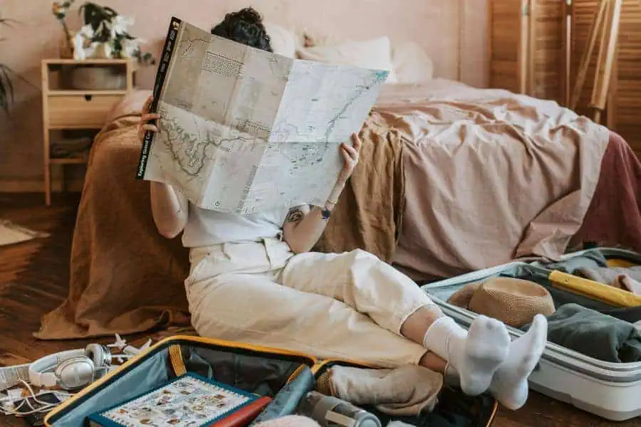 woman sitting on the floor reading a map with open suitcases around, packing for a trip in a cozy bedroom setting